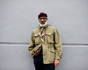 Jason Jules standing against a grey background dressed in beige with a knotted scarf that is patterned. His interests include African American influence on Ivy League style in addition to jazz music. Read the interview at A Collected Man London.